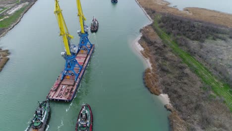 barge being towed through a canal with tugboats and cranes