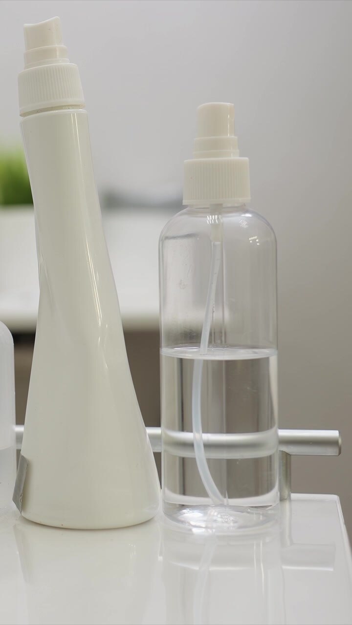 Close up on two bottles one tall with white spray nozzle and another clear container filled with liquid likely for beauty or health treatments. Background is softly blurred emphasizing