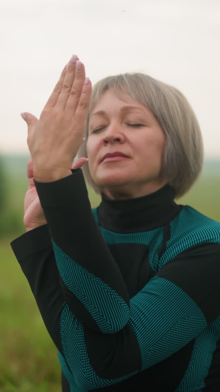 Middle-aged woman in green and black suit practicing yoga outdoors in cow face pose, with eyes closed, focusing on mindfulness and breathing in a serene grassy field under a cloudy sky