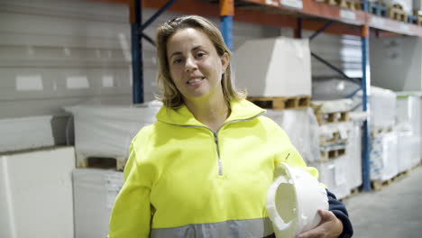 alegre empleada sosteniendo casco, parada en stock y mirando la cámara