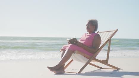 Happy-senior-african-american-woman-sitting-on-deck-chair-and-reading-book-at-beach,-in-slow-motion