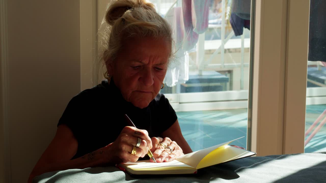 Mezzo busto di una donna anziana concentrata a scrivere sul suo quaderno con una penna stilografica. Scena tranquilla di una persona anziana che scrive un diario a casa vicino alla finestra durante una sessione mattutina silenziosa