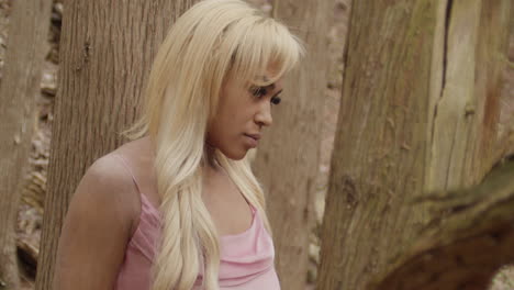 joven mujer de ébano con un vestido rosado. se sienta sola en el bosque, rodeada de árboles. cambia su mirada desde la distancia, a la cámara, luego mira hacia el cielo. cámara de mano a cámara lenta 60 fps.