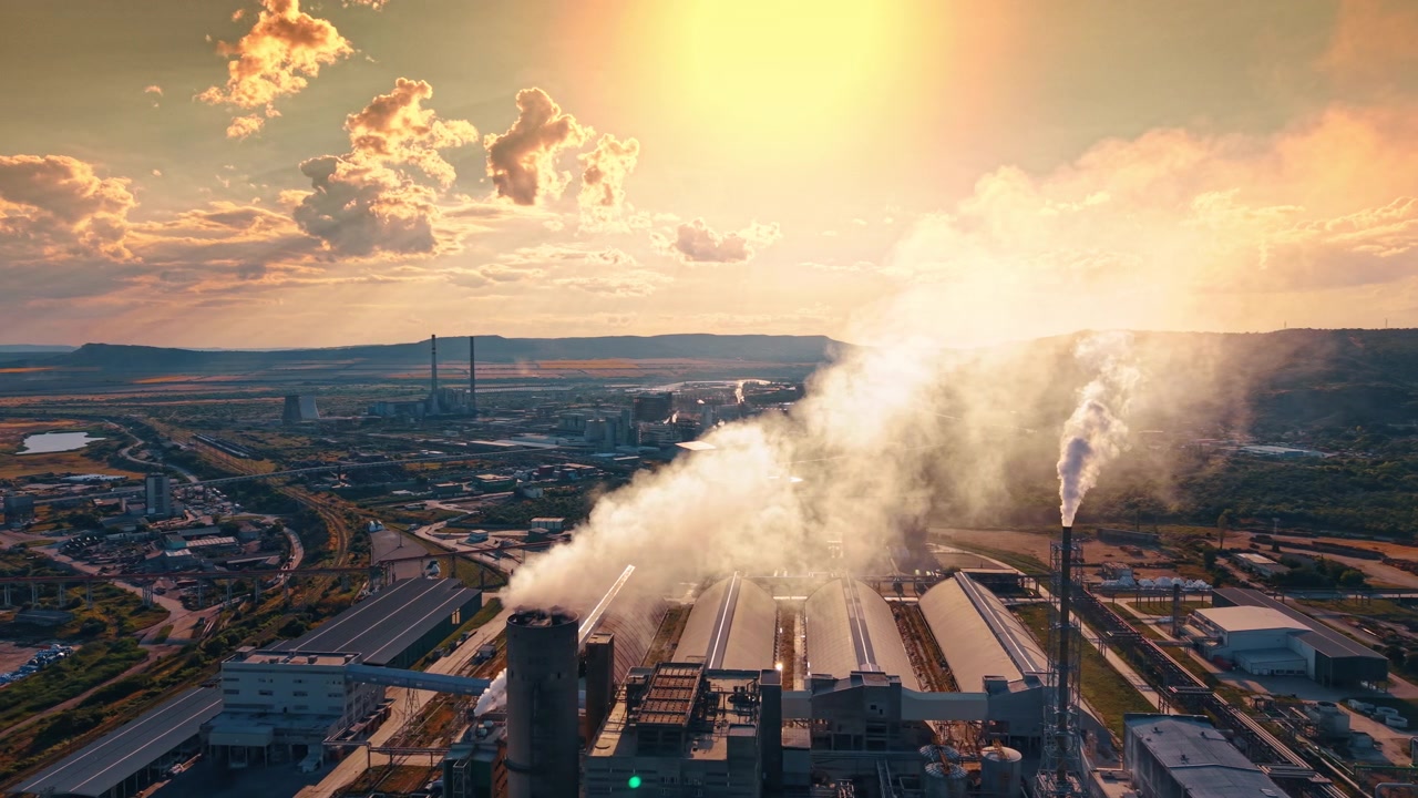 Impianto industriale che emette fumo contro un cielo nuvoloso al tramonto. Forti emissioni si innalzano dalle ciminiere della fabbrica sopra un vasto complesso manifatturiero sotto la calda luce del sole serale.