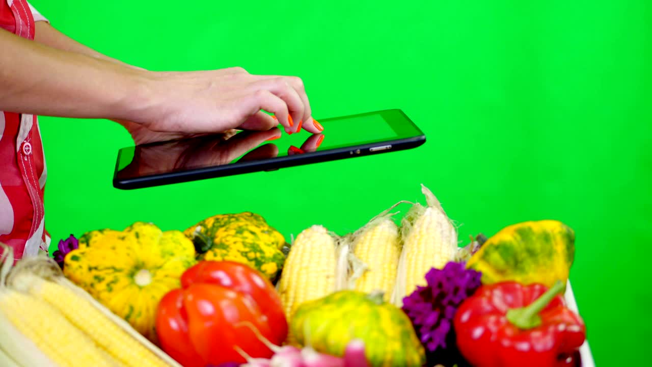 close-up, female hands holding a tablet on Chromakey, green background and a box full of different vegetables, in studio. concept of crop counting, harvest of vegetables