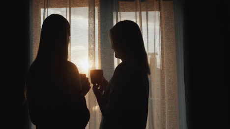 A-woman-with-a-teenage-daughter-stand-at-the-window-at-sunset.-Holding-cups-of-tea