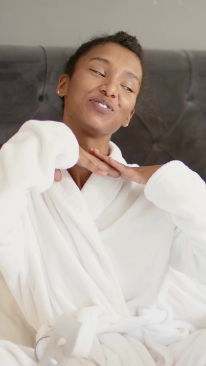 Vertical video: Smiling woman in white robe, camera starting, turning head, touching neck on bed