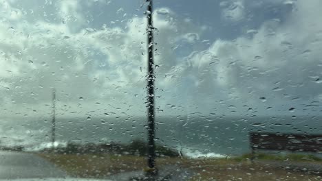 looking through wet windshield at ocean – rain, grass, bench, and cloudy horizon in moody blue coastal landscape