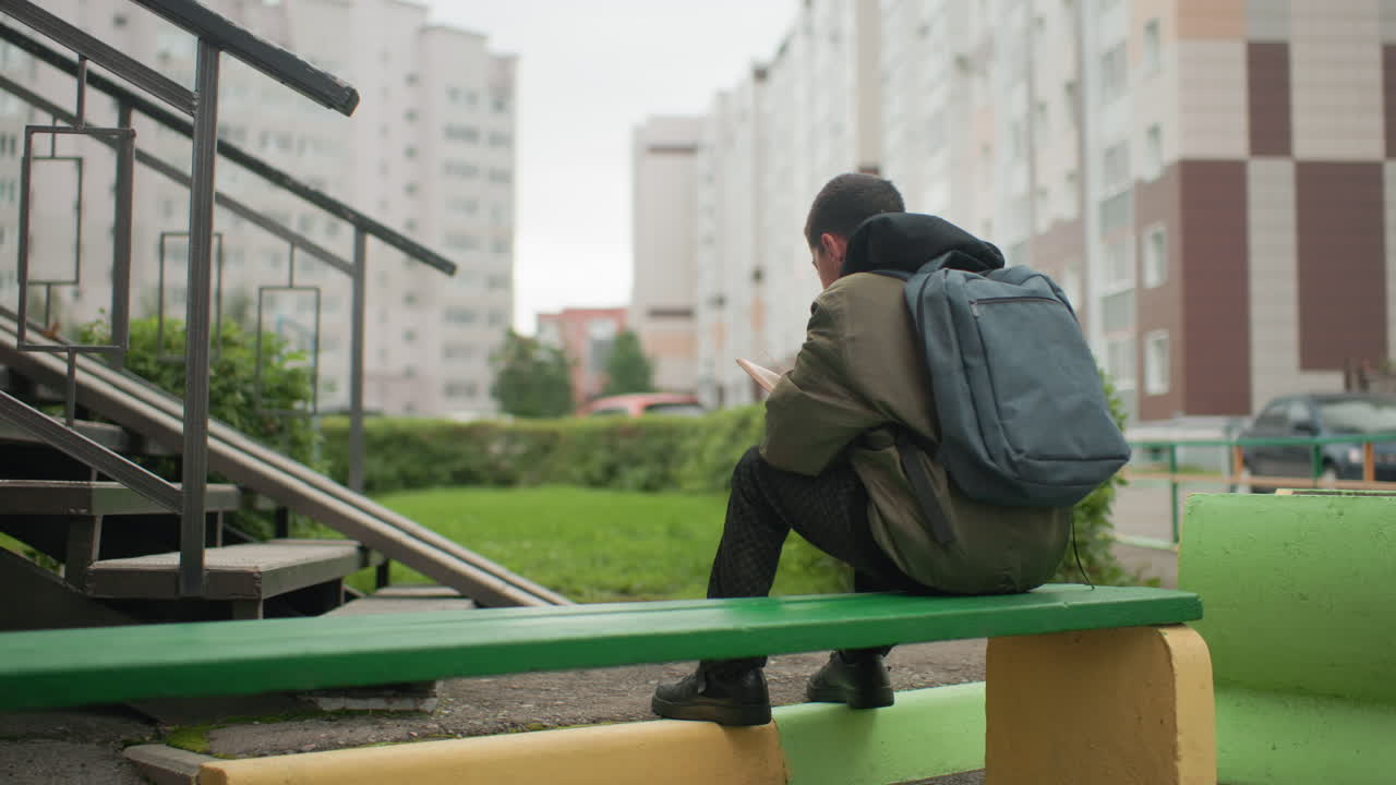 Rear view of young kid wearing backpack seated on green bench reading novel outdoors near apartment building with parked cars and stairway in background