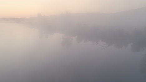 thick fog settles over open fields at dusk, creating a hazy and mysterious landscape, aerial view