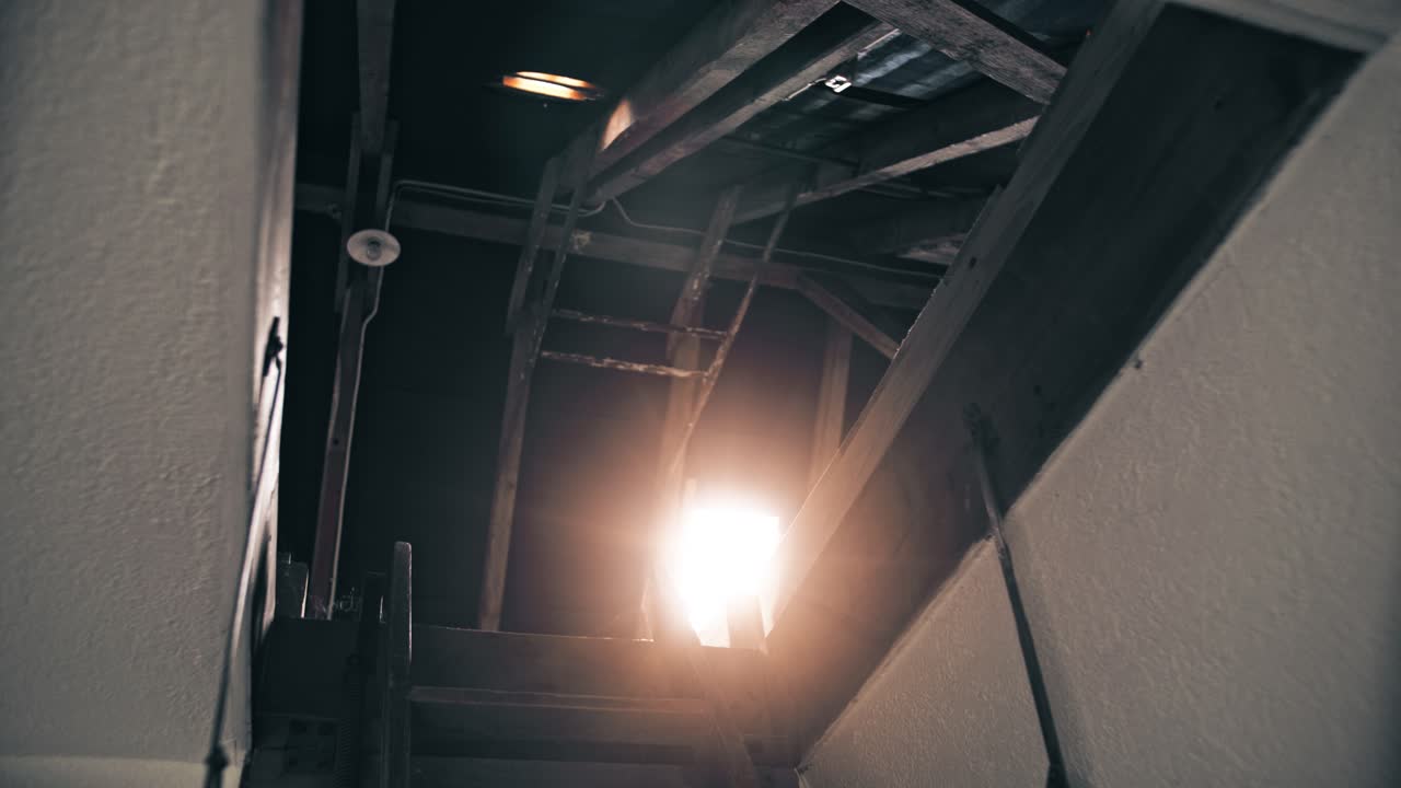 bottom view of an open roof hatch with old and worn staircase and bright orange lens flare shining through the skylight
