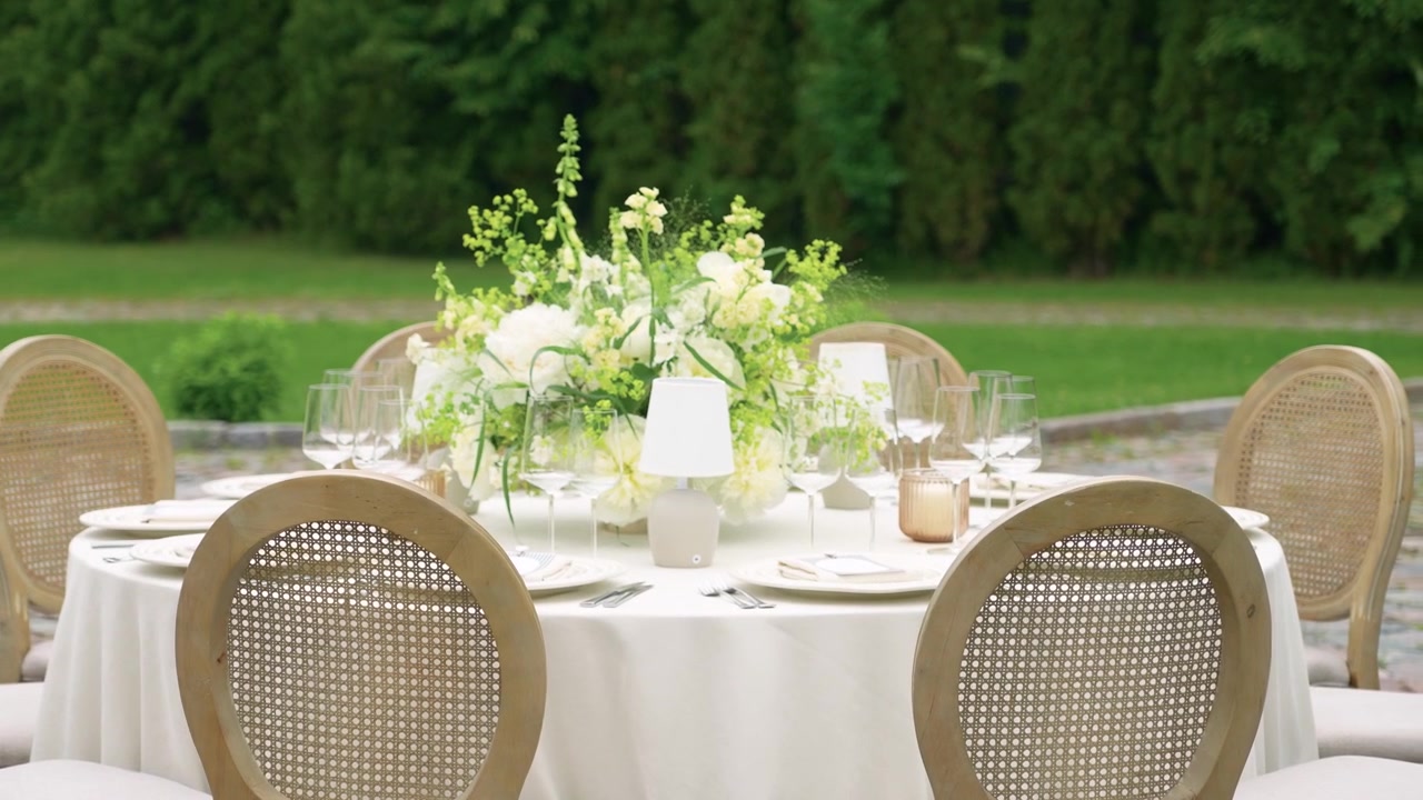 Beautifully decorated round table with cream tablecloth, floral centerpiece, glassware and candles outdoors. Soft daylight, greenery backdrop, and inviting arrangement, perfect for event footage.