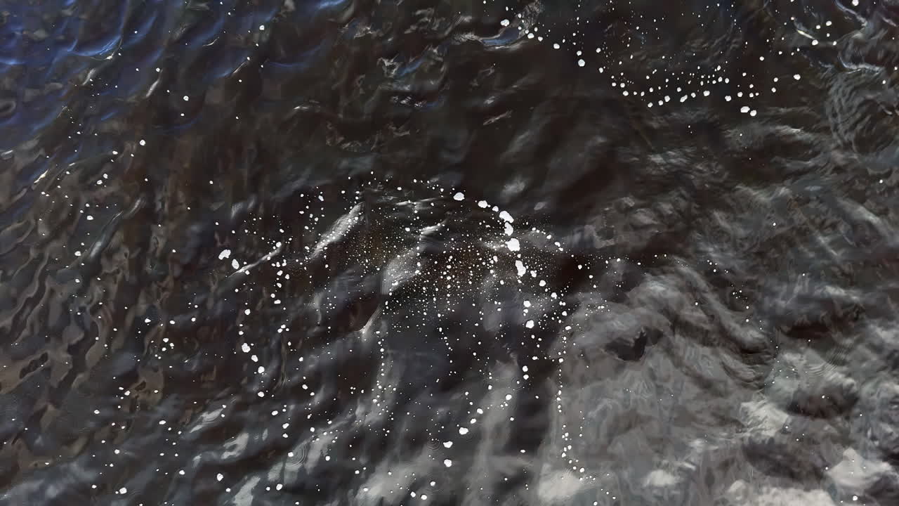 A tight overhead drone shot tracks bright exhalation bubbles as they burst across dark ripples, marking a diver’s position below and suggesting search practice, safety awareness, and quiet tension