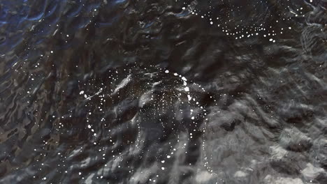 a tight overhead drone shot tracks bright exhalation bubbles as they burst across dark ripples, marking a diver’s position below and suggesting search practice, safety awareness, and quiet tension