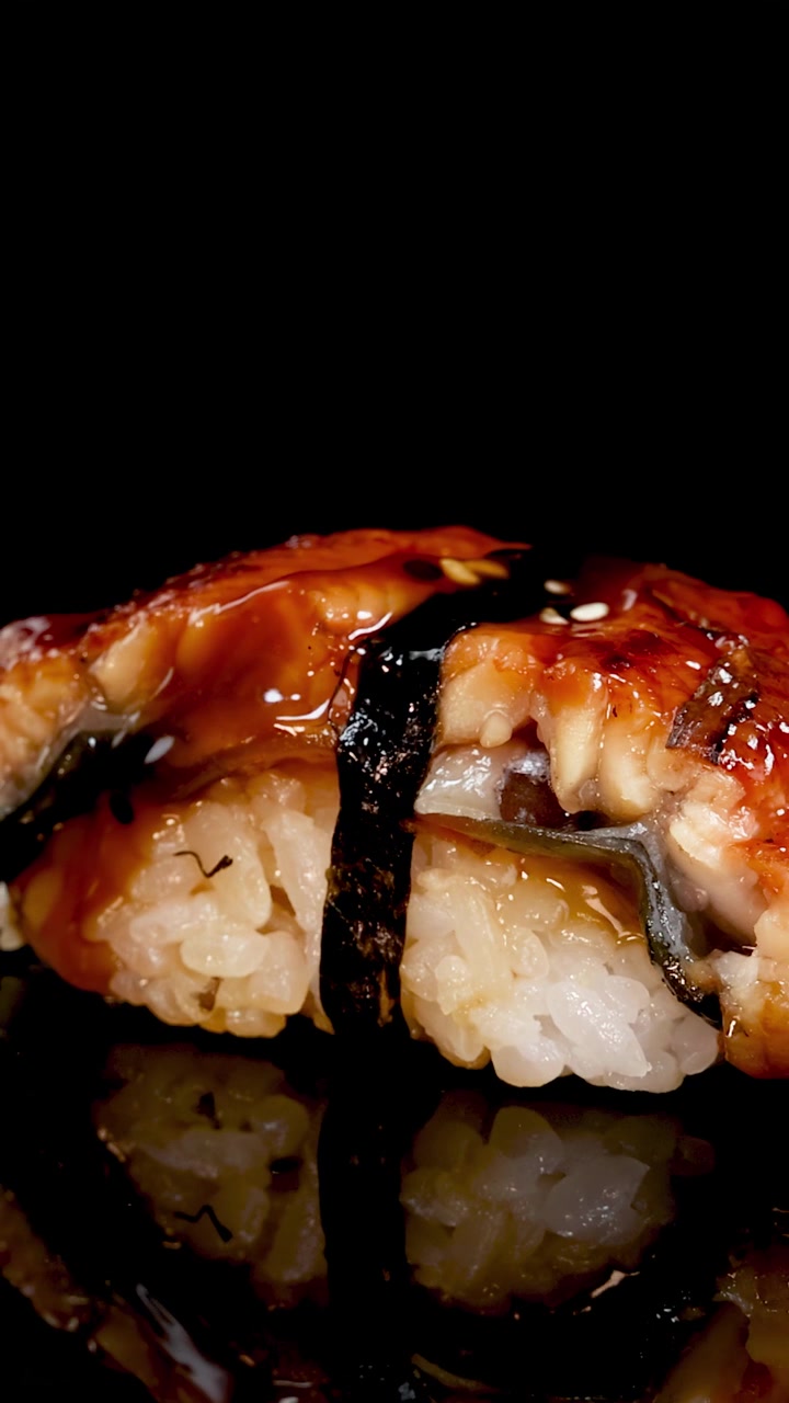 Primo piano di sushi nigiri unagi che ruota lentamente su uno sfondo nero riflettente, con illuminazione da studio drammatica che evidenzia l'anguilla lucida e la salsa.