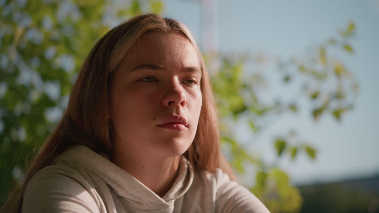 joven en un entorno pacífico al aire libre con los ojos ligeramente cerrados, perdida en los pensamientos, mientras la luz del sol ilumina suavemente su rostro, el fondo muestra hojas verdes borrosas