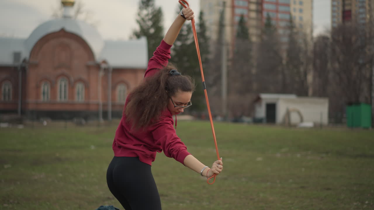 Caucasian Woman Raising Arms In Triumphant Stretch. BackFacing Celebratory Pose In Open Park, Relaxed Shoulders, Long Hair, Distant Buildings And Uplifted Mood After Focused Session