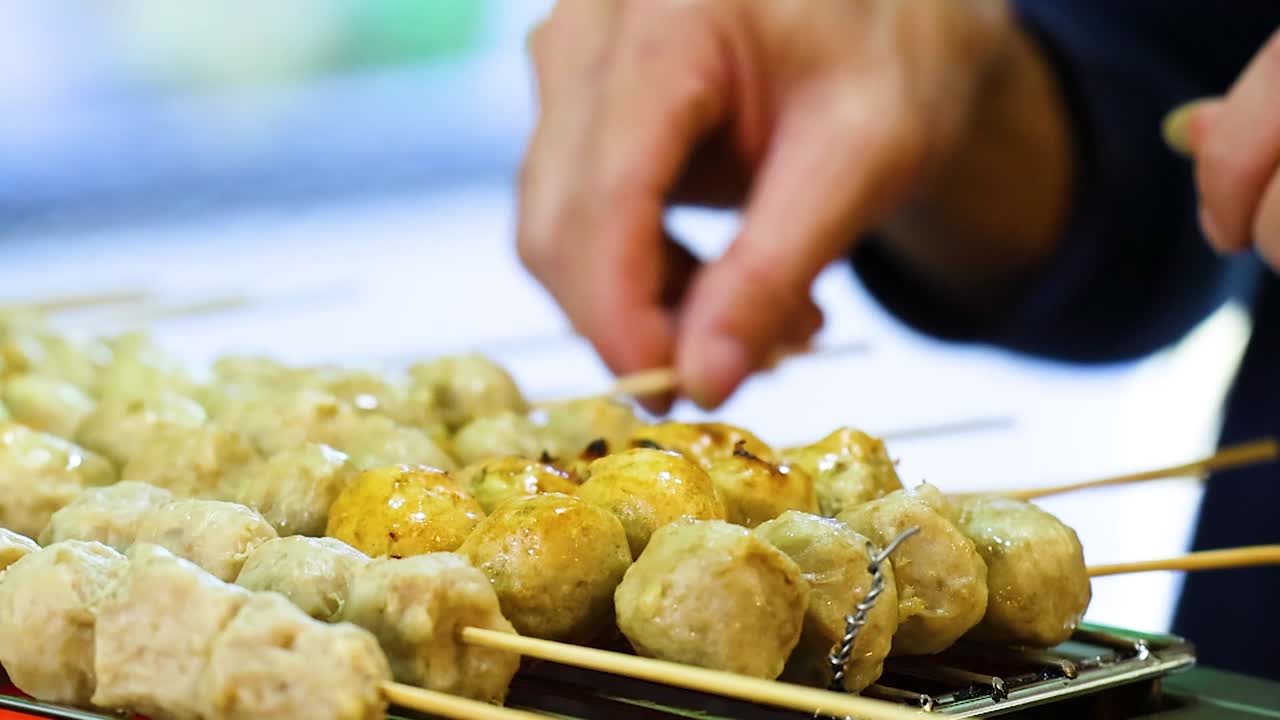 Hands grilling skewered meatballs at a market stall