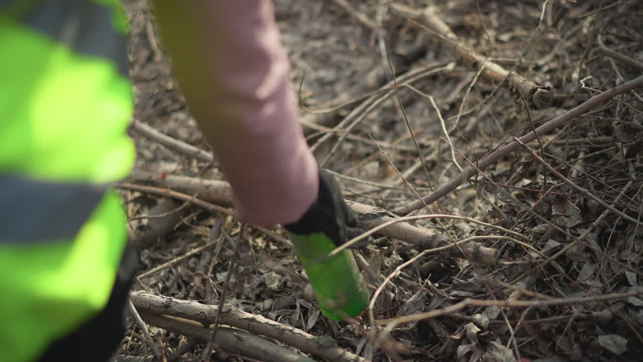 Volunteer wearing high visibility vest and black gloves reaching into pile of dry leaves and branches to pick up hidden litter during outdoor cleanup, removing waste to protect environment