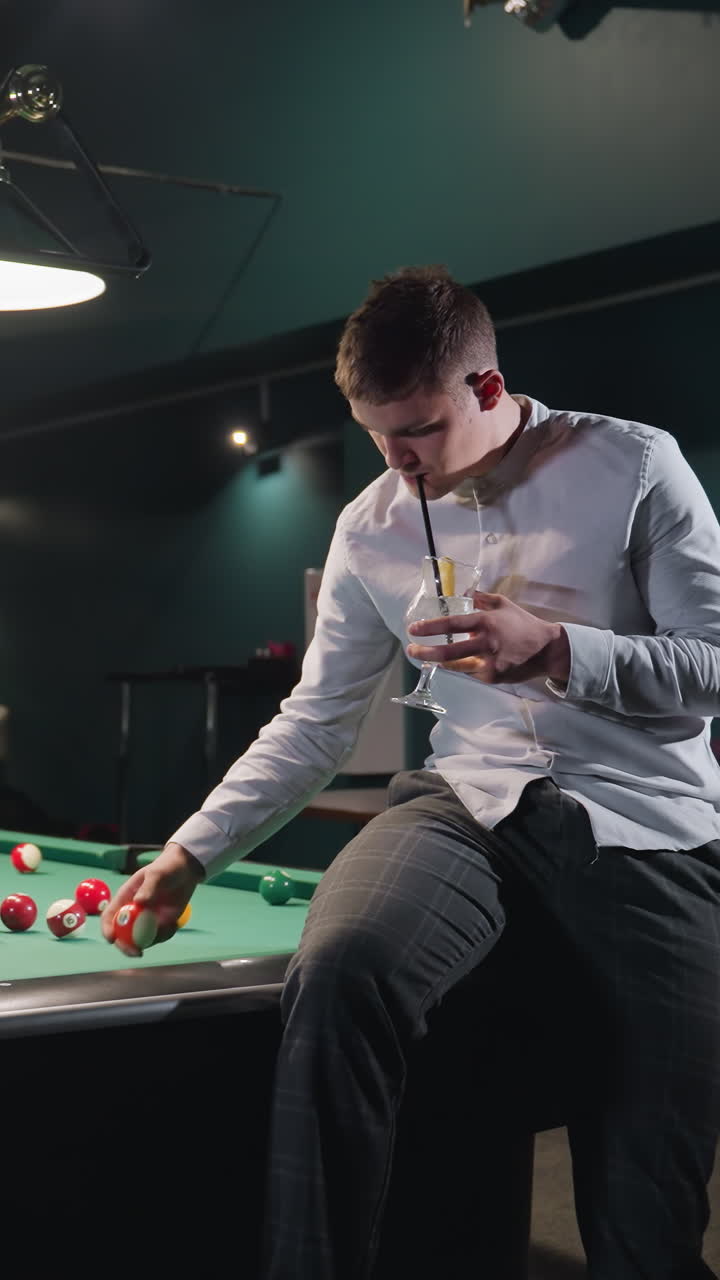 un deportista con camisa blanca y pantalones grises se sienta en una mesa de billar verde, bebiendo una bebida de limón con paja negra en una sala de billar débilmente iluminada.