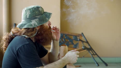 homeless man lighting up marijuana or tobacco cigarette and drinking beer can outdoors near broken bench