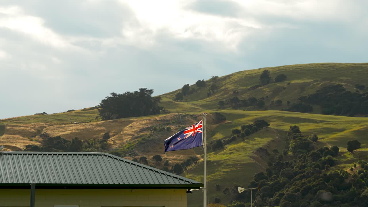 New Zealand flag next to building waving in slomo with lush hills in background
