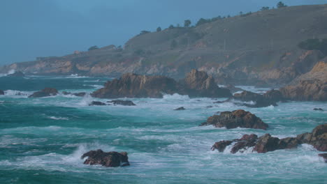 Painters-Point,-California---wide-shot-pan-of-rough-surf-with-Highway-1-on-top-of-hill