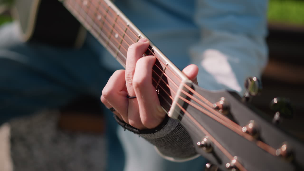 dita di un chitarrista bianco che formano un accordo aperto su una chitarra acustica all'aperto, primo piano della tastiera e delle corde, camicia di jeans, tecnica di plettraggio energica, prova su una panchina in strada, luce solare calda, concentrato