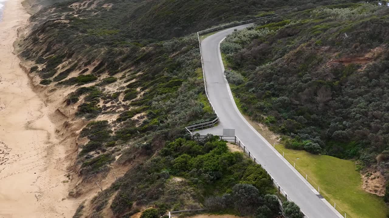 Drone glides above winding coastal road, modern house, sandy beach, and lush greenery in daylight