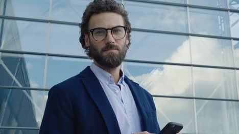 young confident professor in glasses looking to the camera confidently. portrait of young caucasian man with stylish hair and look. using his smartphone. social network. work outside. success. people