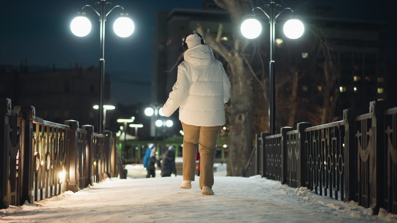 Female artist spinning joyfully on snow covered park walkway at night under bright lamp posts with distant figures playing and walking, creating festive winter ambiance and dynamic urban energy