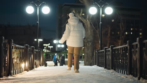 female artist spinning joyfully on snow covered park walkway at night under bright lamp posts with distant figures playing and walking, creating festive winter ambiance and dynamic urban energy