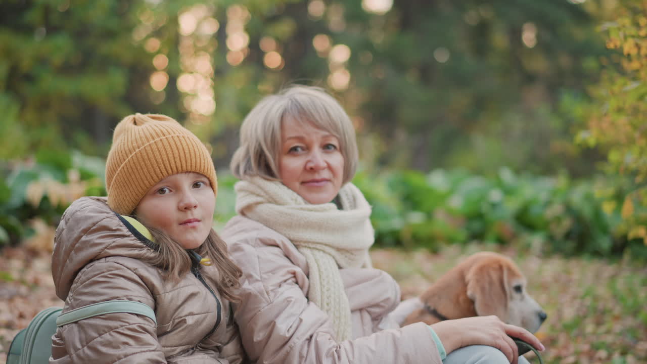 mom and daughter wearing warm jackets look calmly into camera while sitting on autumn leaf strewn ground with dog resting beside them in peaceful green woodland setting during soft daylight