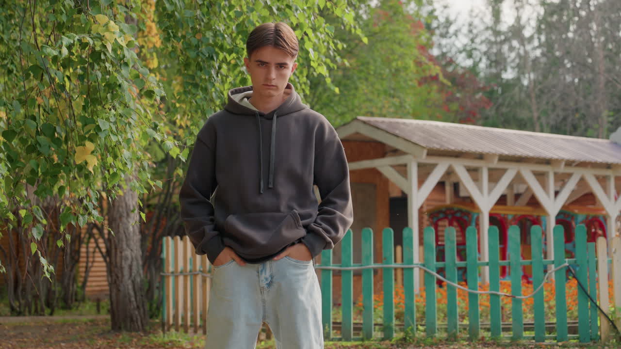 Kaukasischer Teenager steht neben einem Zaun im Garten, Ganzkörperaufnahme mit Lattenzaun und Hütte dahinter, lässiger Hoodie und Jeans, selbstbewusste Haltung, Herbstlaub unter den Füßen, Rollen deuten auf Straße hin