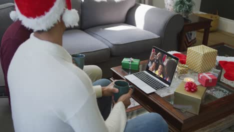 Padre-E-Hijo-Birraciales-Con-Gorros-De-Papá-Noel-Usando-Una-Computadora-Portátil-Para-Una-Videollamada-Navideña-Con-La-Familia-En-La-Pantalla