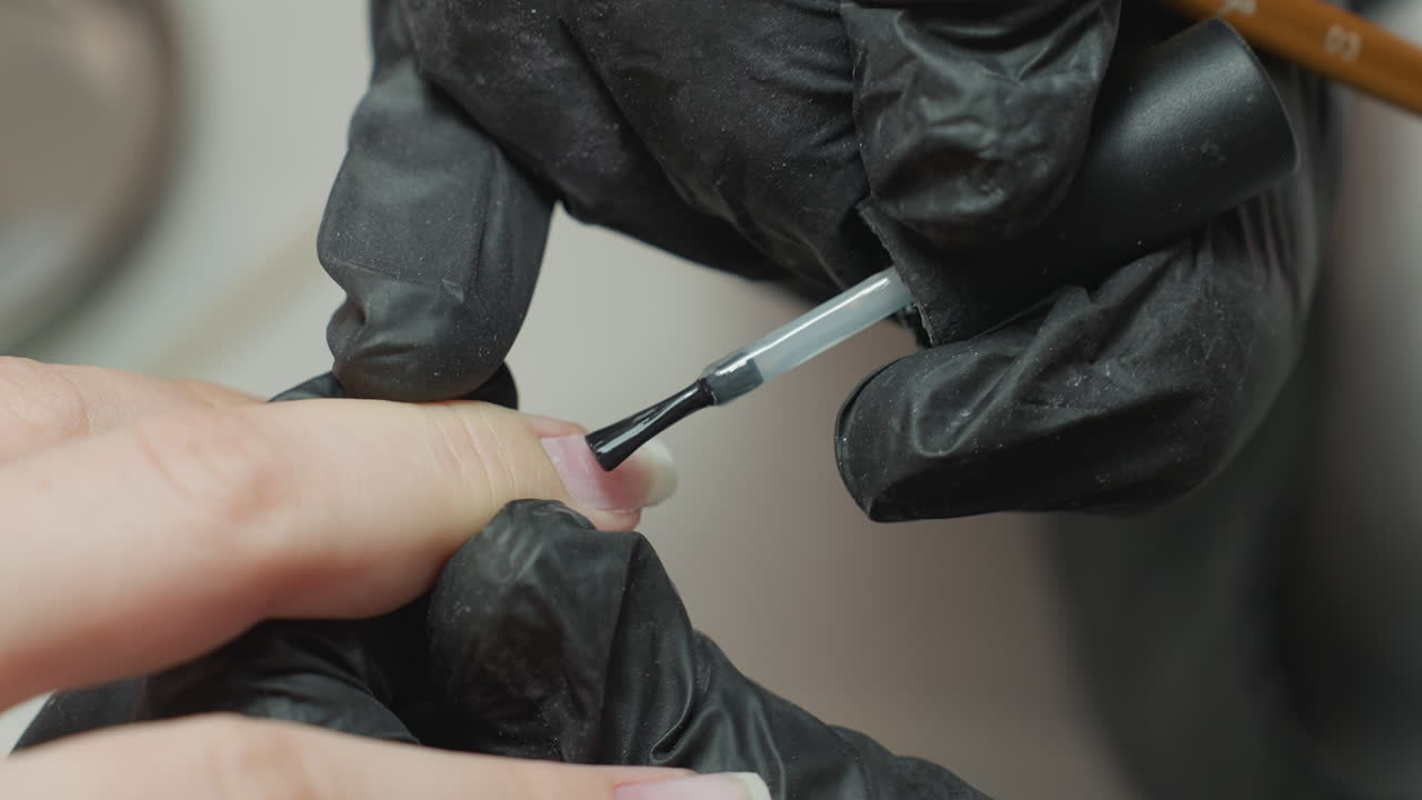 Close-up of nail technician wearing dusty black glove gently applying clear nail hardener to client's fingernail with precise brush strokes during manicure session in professional beauty salon