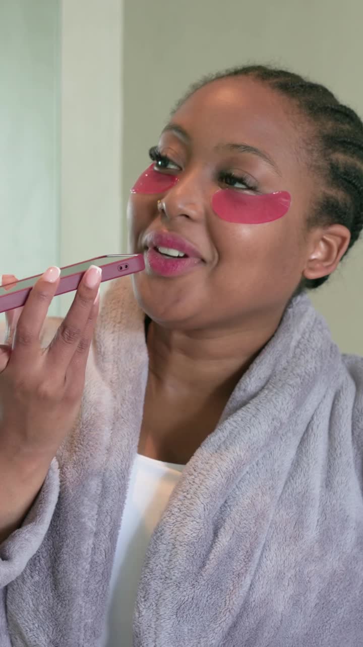 Vertical video: Glancing at pink phone, lady in robe at sink raising it, speaking, showing patches