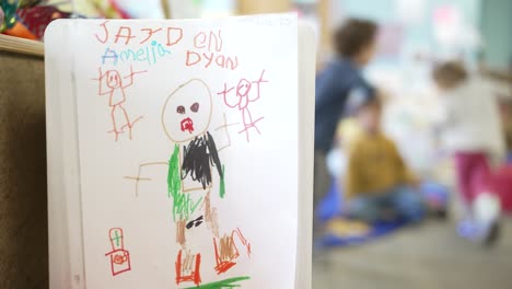 a preschooler's drawing hung up with unidentifiable children playing in background