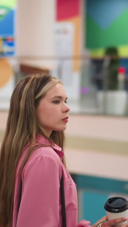 woman in pink dress holding coffee cup and shopping bags while standing on an escalator as it descends in a brightly lit mall, colorful wall designs and glass railings can be seen in the background