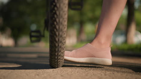 close-up of individual legs in pink sneakers kicking a bicycle tire to check air pressure, lush greenery and trees fill the background, with an indistinct figure moving