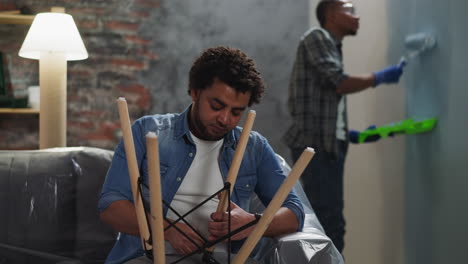 Black-man-assembles-chair-and-colleague-paints-wall-in-room