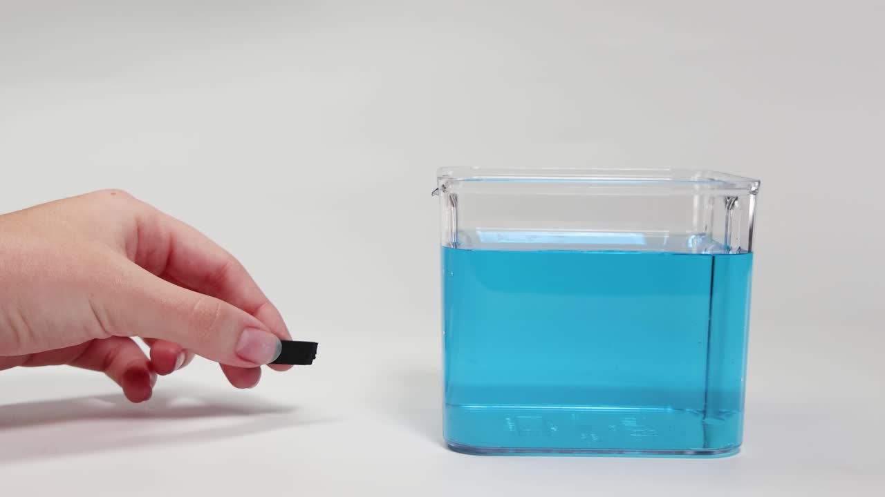 A hand places a rubber foam block in water, demonstrating buoyancy. Clear lighting and simple composition highlight the scientific process