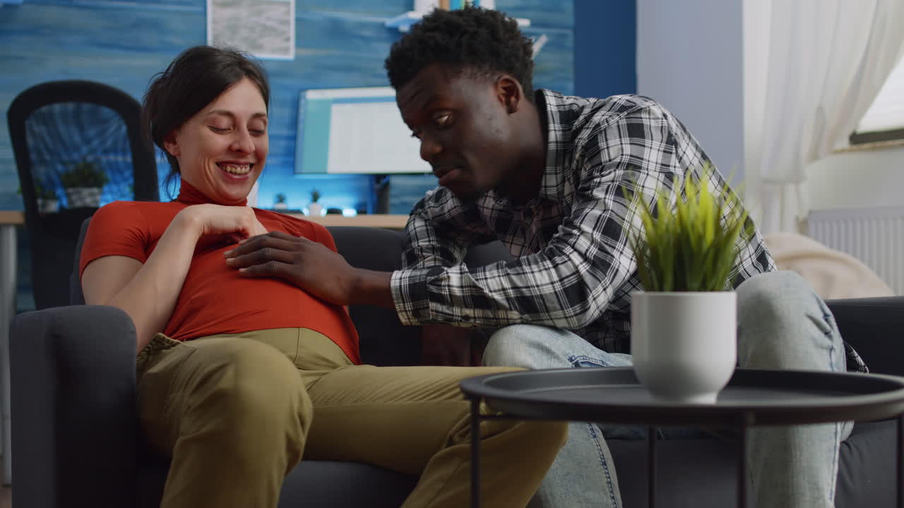 Young interracial couple expecting child sitting on sofa
