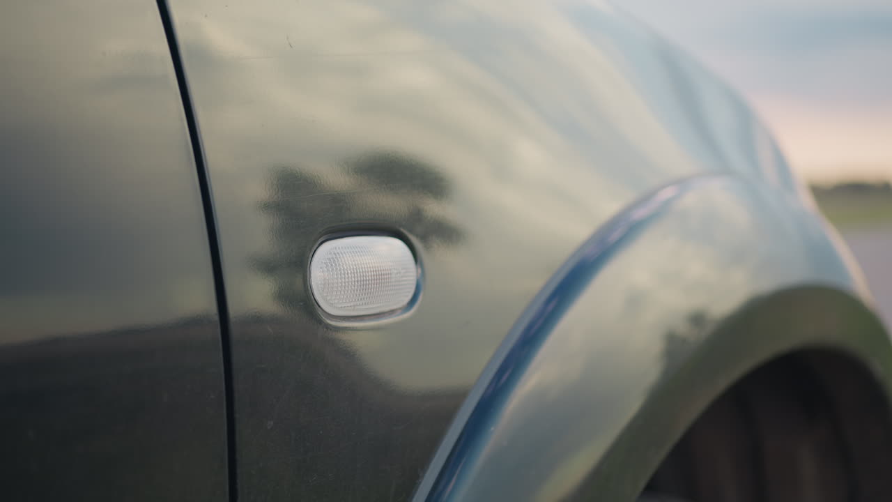 Primo piano dell'indicatore di direzione laterale dell'auto che lampeggia delicatamente con riflessi di cielo soffice e silhouette di alberi su una superficie lucida e scura, mentre un campo aperto in lontananza sfuma in una leggera sfocatura sotto una luce tranquilla