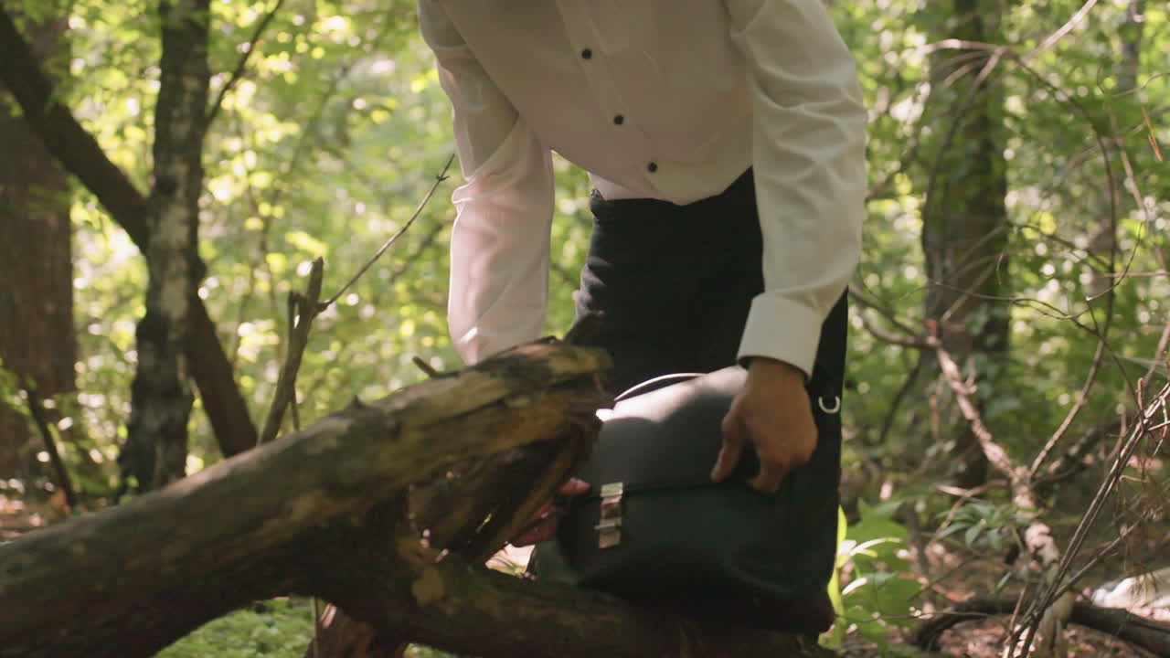 Scientific researcher in white shirt putting white coat inside backpack bag while standing in forest, surrounded by green trees and sunlight, preparing for outdoor activity with focus and purpose