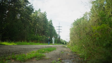 rear view of little boy carrying black backpack walking with sister along cracked paved road surrounded by green forest and power lines under bright sky, bonding during calm outdoor journey together