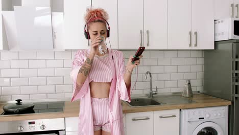 happy young woman with pink hair dancing in kitchen wearing pink pajamas and listening to music with headphones