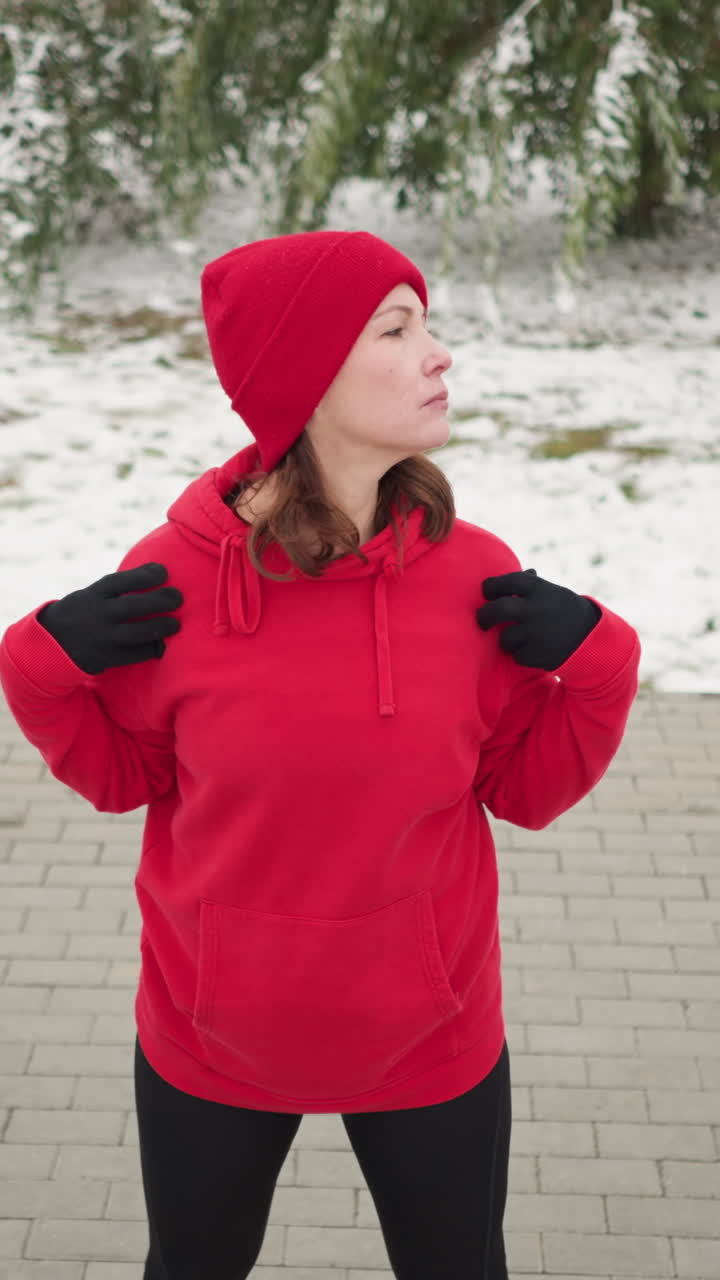 어깨에 손을 은 빨간 후디를 입은 여인이 눈이 내린 야외 환경에서 머리를 천천히 왼쪽으로 돌리고, 얼음 나무, 벤치에 가방, 그리고 눈으로 인 땅