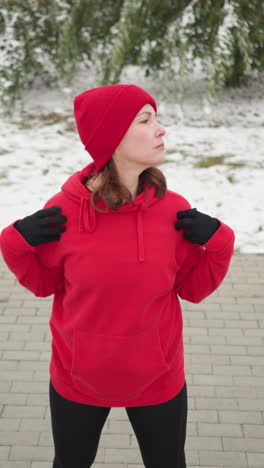 dama con capucha roja con las manos colocadas sobre los hombros gira la cabeza lentamente hacia la izquierda en un entorno al aire libre nevado con árbol helado, bolsa en el banco y tierra cubierta de nieve