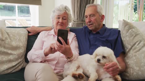 Una-Pareja-Caucásica-Mayor-Sentada-En-Un-Sofá-Con-Su-Perro-Mascota-Usando-Un-Teléfono-Inteligente-En-La-Sala-De-Estar-En-M-Lento